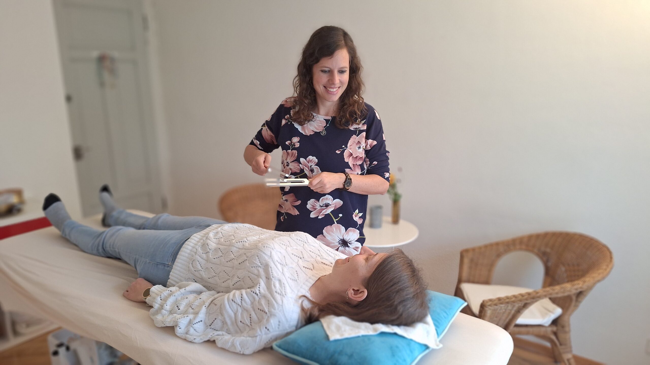 Therapeutin hält Stimmgabeln über einer auf einer Liege liegenden Patientin in einem hellen Behandlungsraum.