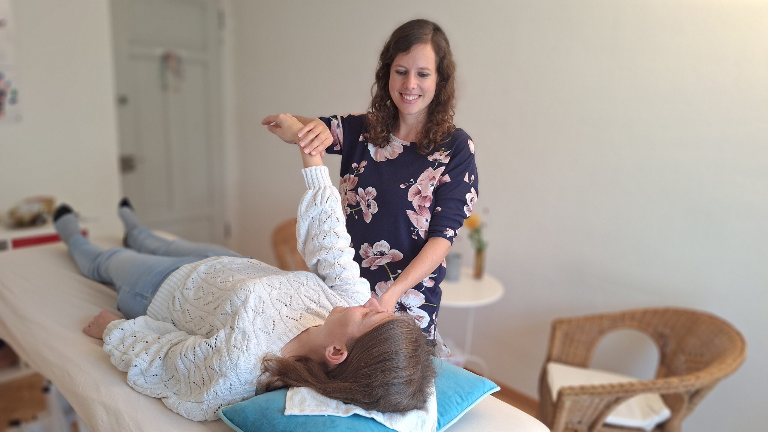 Therapeutin prüft den Arm einer auf einer Liege liegenden Patientin in einem hellen Behandlungsraum.