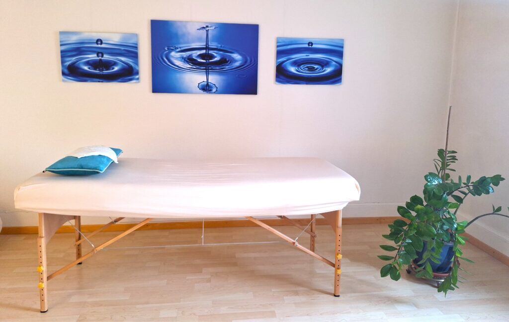 Heller Behandlungsraum mit einer Massageliege, Wandbildern von Wassertropfen und einer grünen Pflanze.